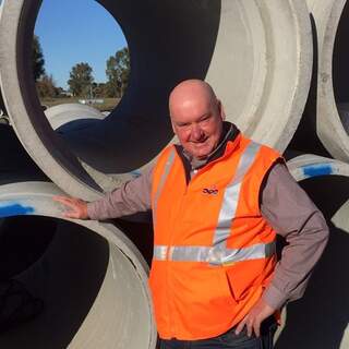 Photograph of Tony Corbett wearing high vis clothing on a project site standing in front of cement drainage pipes